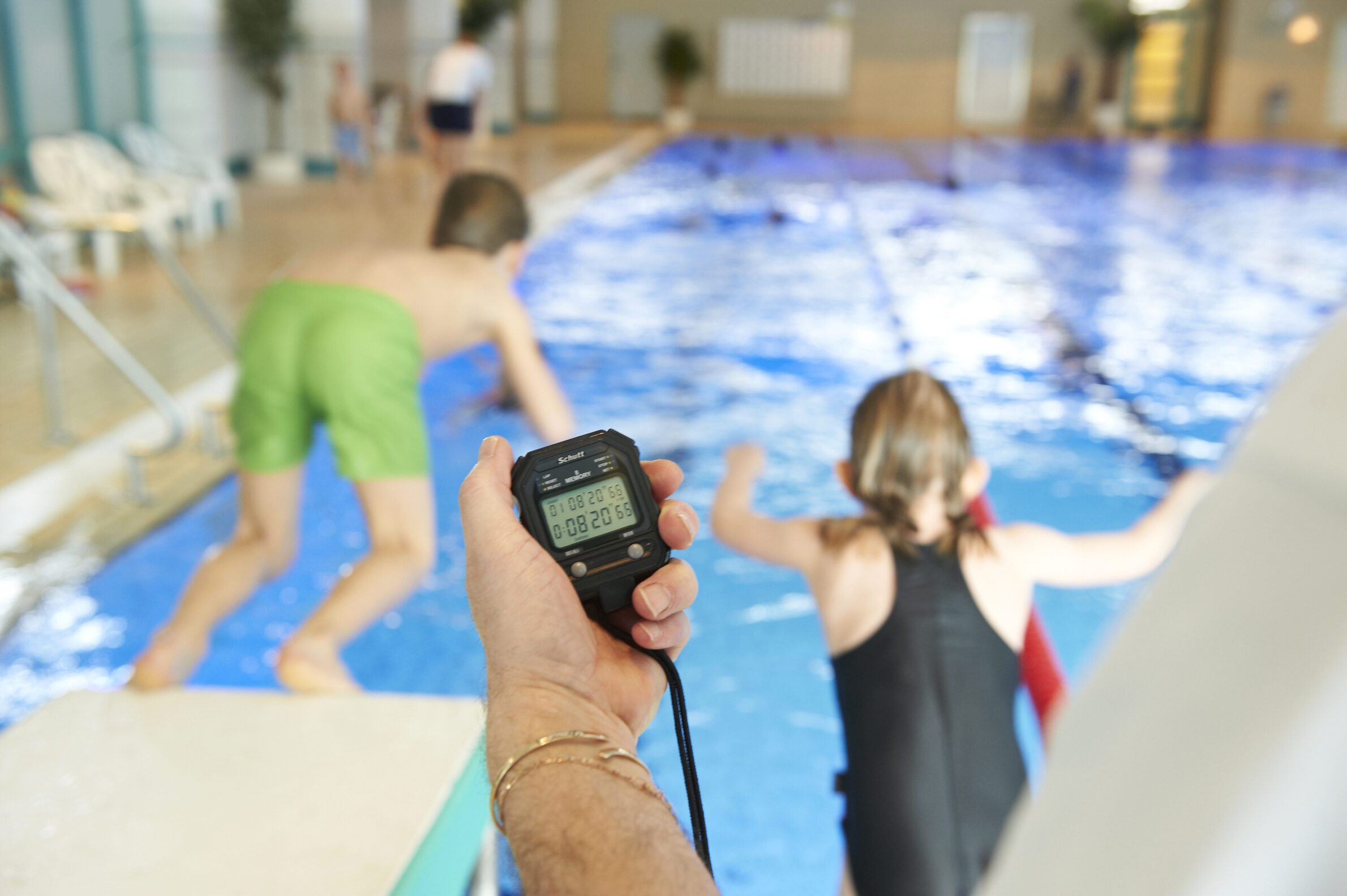 Stoppuhr im Vordergrund zwei Kinder springen in Schwimmerbecken